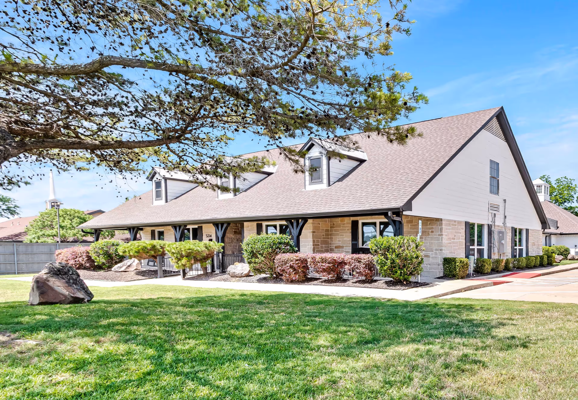 Exterior view of a single-story assisted living facility building with a sloped roof, dormer windows, and a well-maintained lawn with bushes and rocks in front. A large tree branch extends over the lawn, and the sky is clear and blue.
