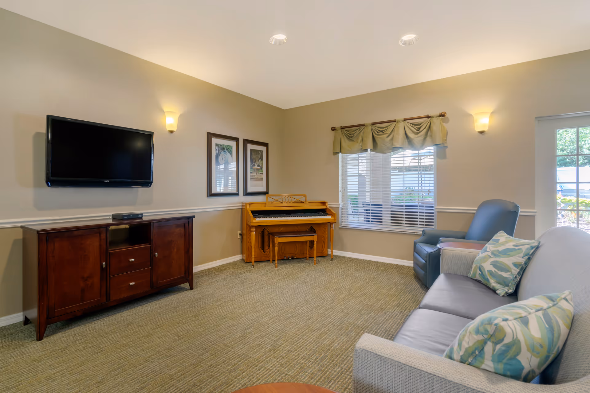 A bright common living room with a wall-mounted TV, wooden cabinet, small piano, sofa and armchair by a window.