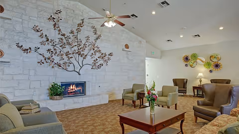 A cozy living room area with a stone wall featuring a built-in fireplace and a decorative metal tree sculpture above it. The room has several upholstered chairs and sofas arranged around a wooden coffee table with a flower vase. The carpet is patterned, and the ceiling has recessed lighting and a ceiling fan. There are decorative wall hangings and a table lamp in the background.