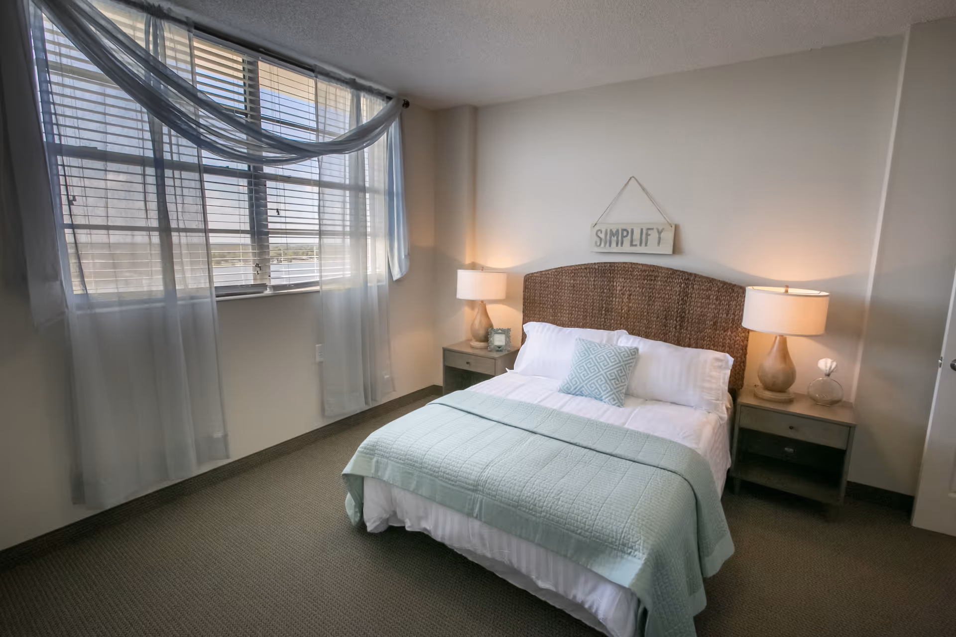 A bright bedroom with a bed, wicker headboard, two bedside lamps, and a large window with sheer curtains.