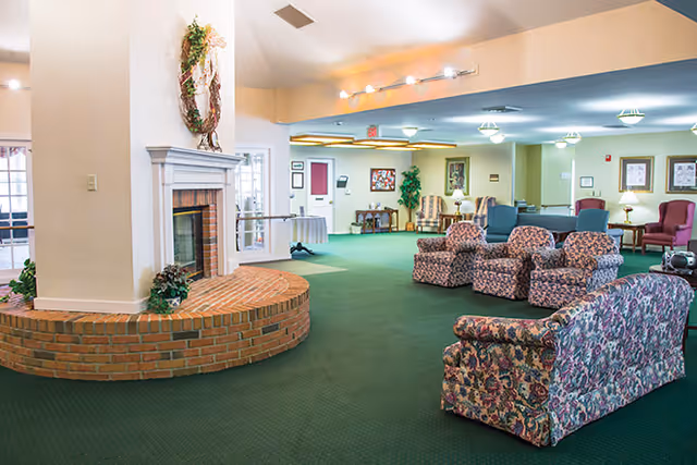 A spacious senior living facility common area with green carpet, floral patterned armchairs and sofa, a brick fireplace with a decorative wreath above it, and soft lighting from ceiling fixtures. The room has a cozy and welcoming atmosphere with framed artwork and plants in the background.