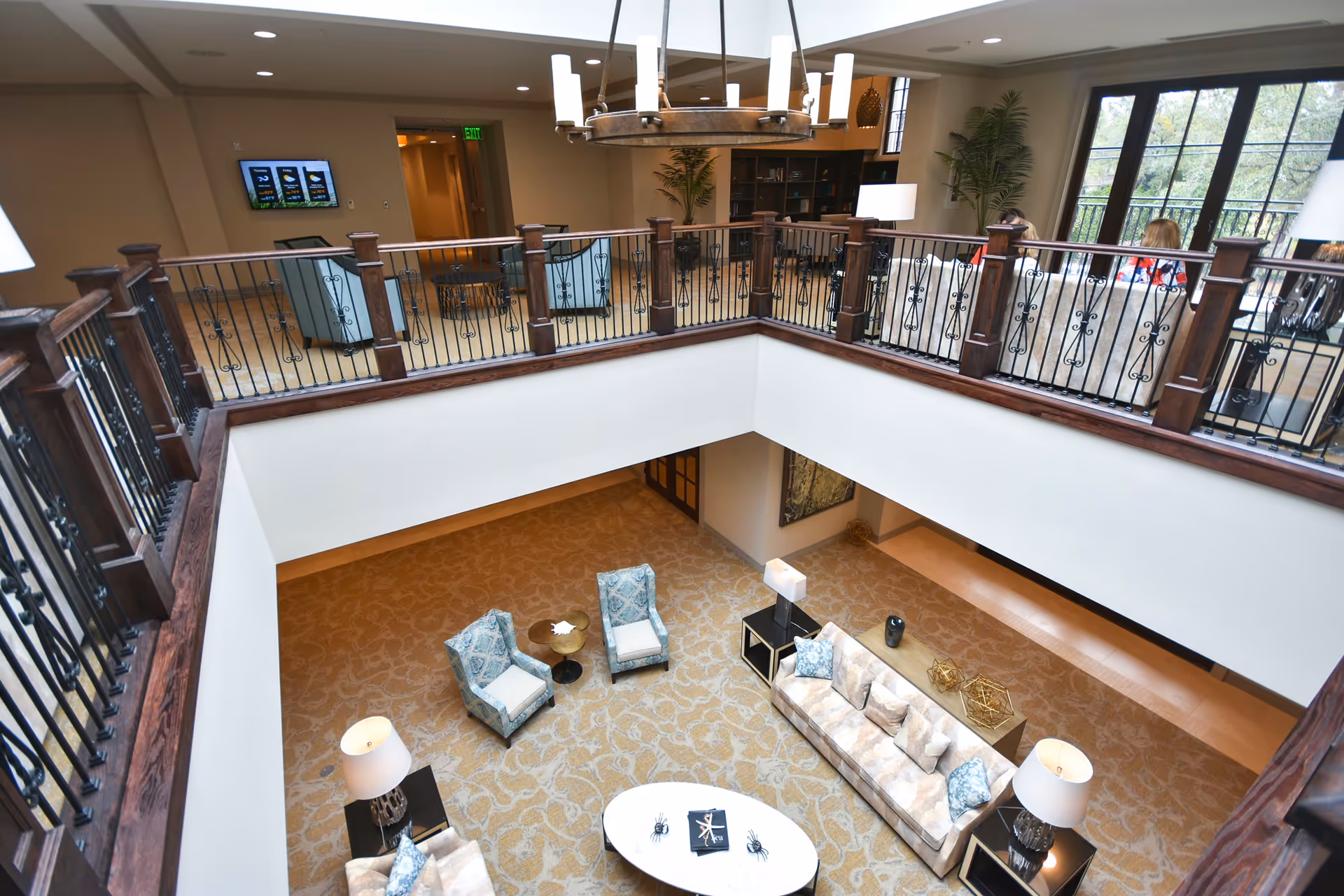 View from an upper floor overlooking a spacious living room area with a beige patterned carpet, a beige sofa with blue and beige cushions, two blue patterned armchairs, a white oval coffee table, and two side tables with lamps. The upper floor has a wooden railing with black iron balusters, and there are additional seating areas and large windows letting in natural light.