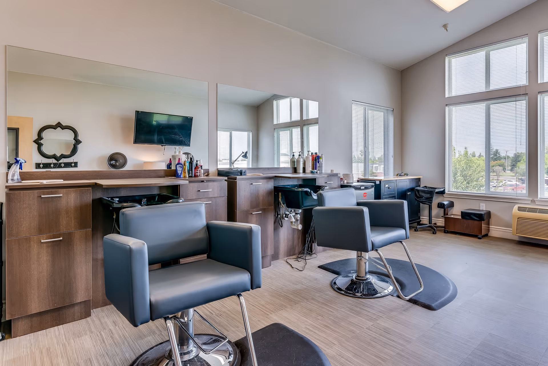 A bright and modern hair salon area with two gray salon chairs in front of large mirrors mounted on a wall with wooden cabinetry. The room has large windows letting in natural light, and various hair care products are placed on the counters.