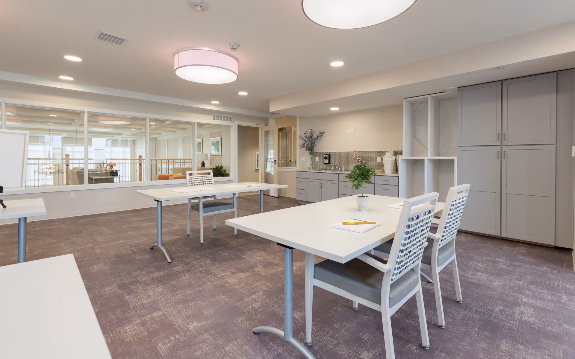 A bright, modern activity or meeting room with white tables and chairs arranged in pairs. The room features a purple carpet, gray cabinetry with a small sink, and decorative plants on the tables. Large windows look into an adjacent lounge area with seating and artwork on the walls.