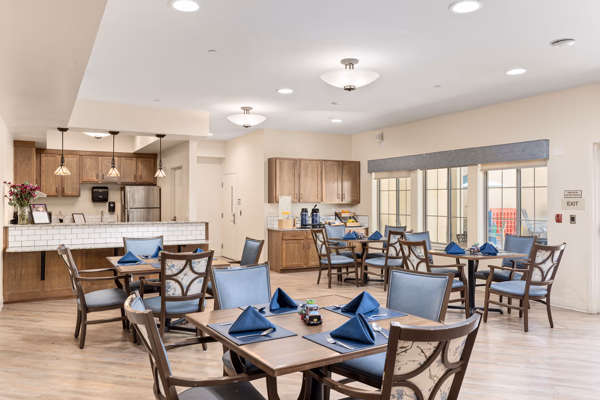 A bright dining room with wooden tables and chairs set with blue napkins and silverware. The room features large windows with a view outside, wooden cabinetry, and a kitchen area with pendant lights and a stainless steel refrigerator.