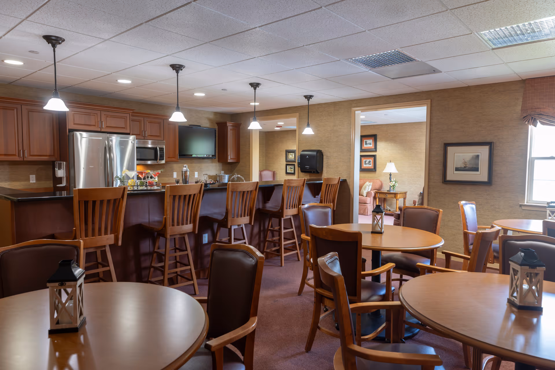 A cozy dining area in an assisted living facility featuring round wooden tables with chairs, each table decorated with a small lantern. In the background, there is a kitchen area with wooden cabinets, stainless steel refrigerator, microwave, and a counter with high wooden chairs. Pendant lights hang from the ceiling, and through an open doorway, a living room with a sofa and framed pictures on the wall is visible.