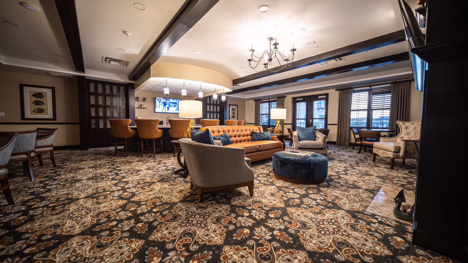 A spacious and elegantly decorated living room area with a patterned carpet, a brown leather sofa, several upholstered chairs, a blue round ottoman, and two table lamps. The room features large windows with blinds and curtains, a chandelier on the ceiling, and a bar area with high chairs and a TV mounted on the wall.