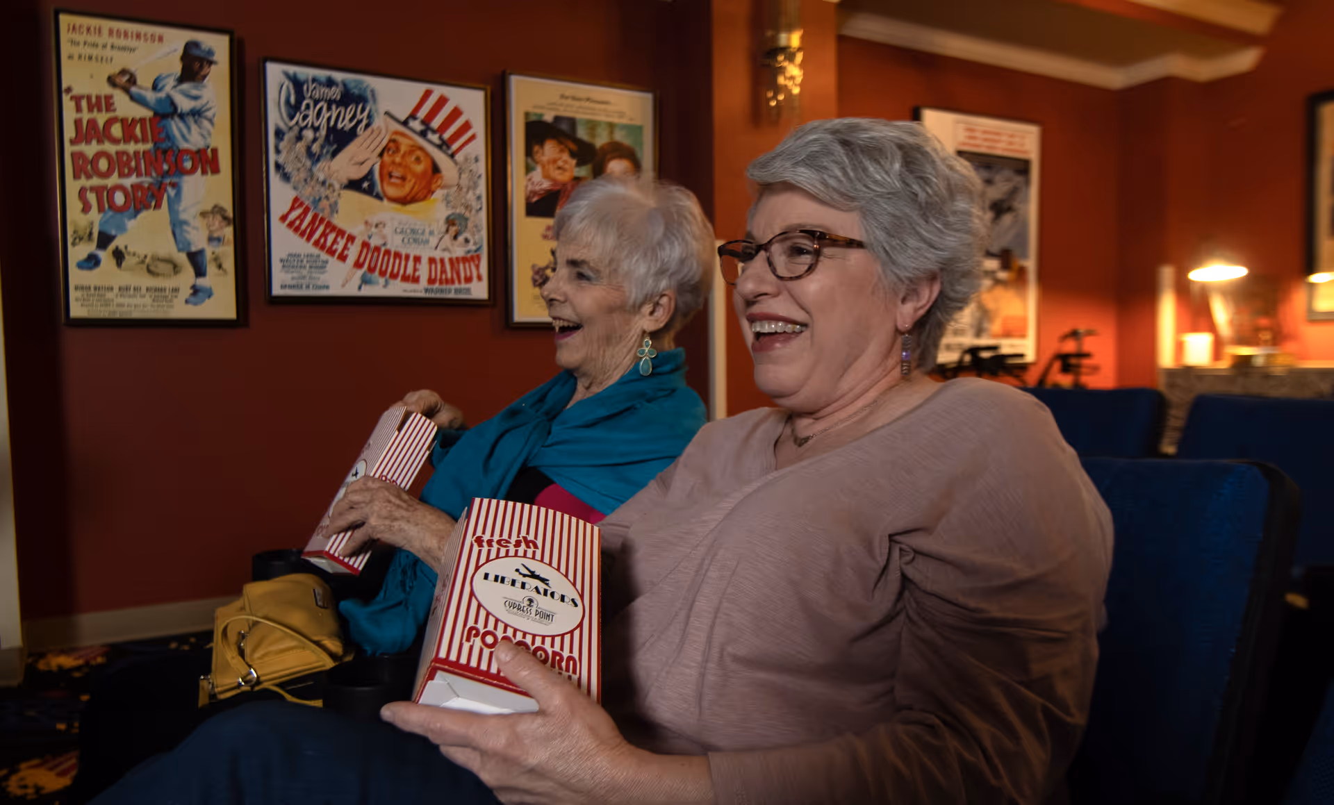 Two elderly women sitting in a theater room, smiling and holding striped popcorn boxes. The walls behind them are decorated with vintage movie posters including The Jackie Robinson Story and Yankee Doodle Dandy.