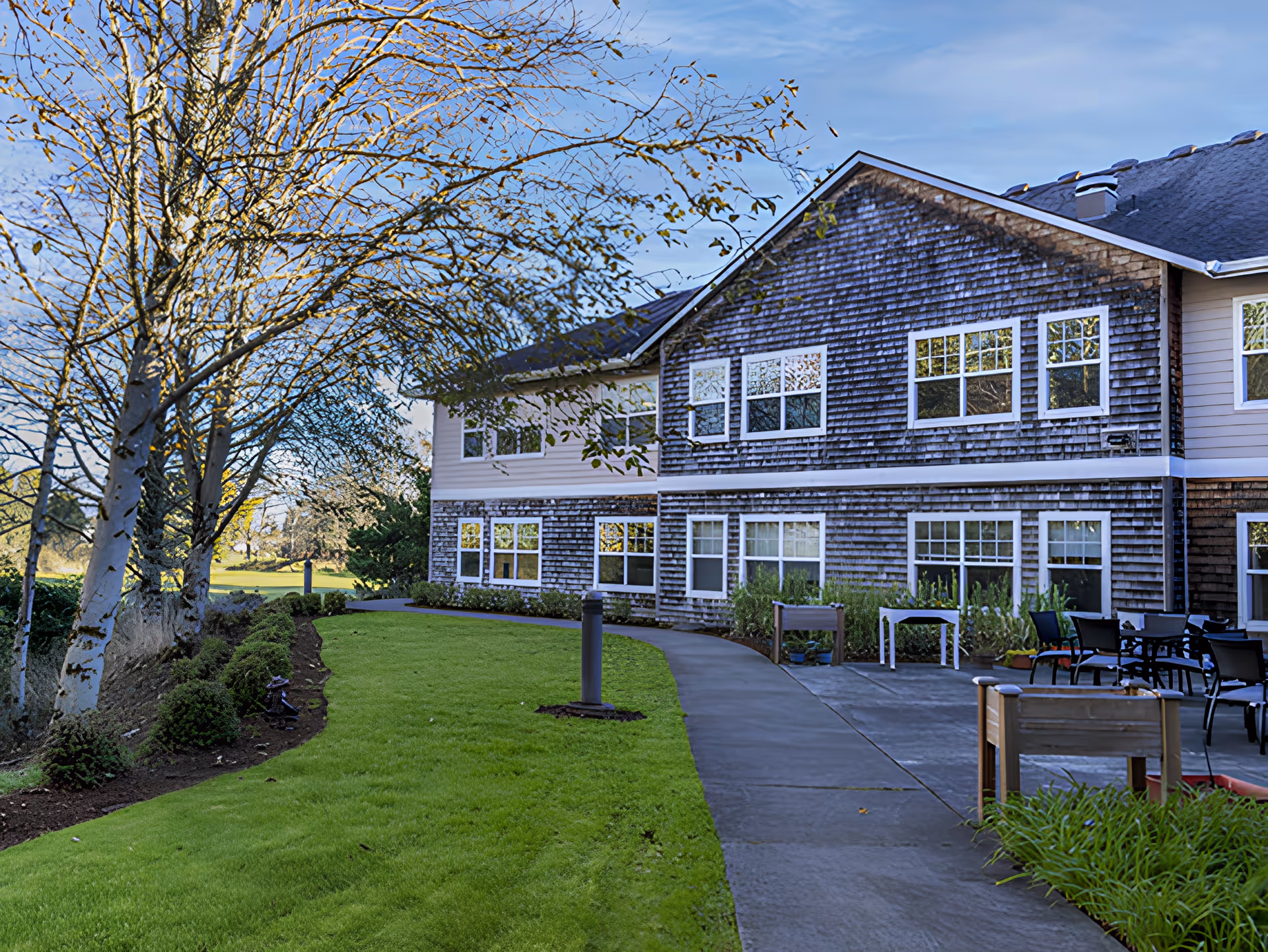 Outdoor view of a senior living facility building with wooden siding and multiple windows. A paved walkway curves through a well-maintained green lawn with small bushes and trees. There are outdoor tables and chairs on a patio area near the building.