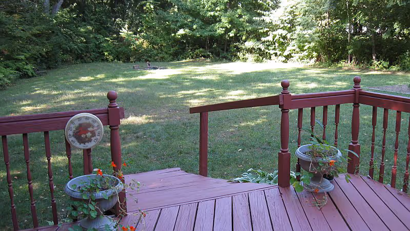 View from a wooden deck with red railing overlooking a grassy yard surrounded by trees. Two planters with flowers are placed on the deck, and a round thermometer is attached to the railing.