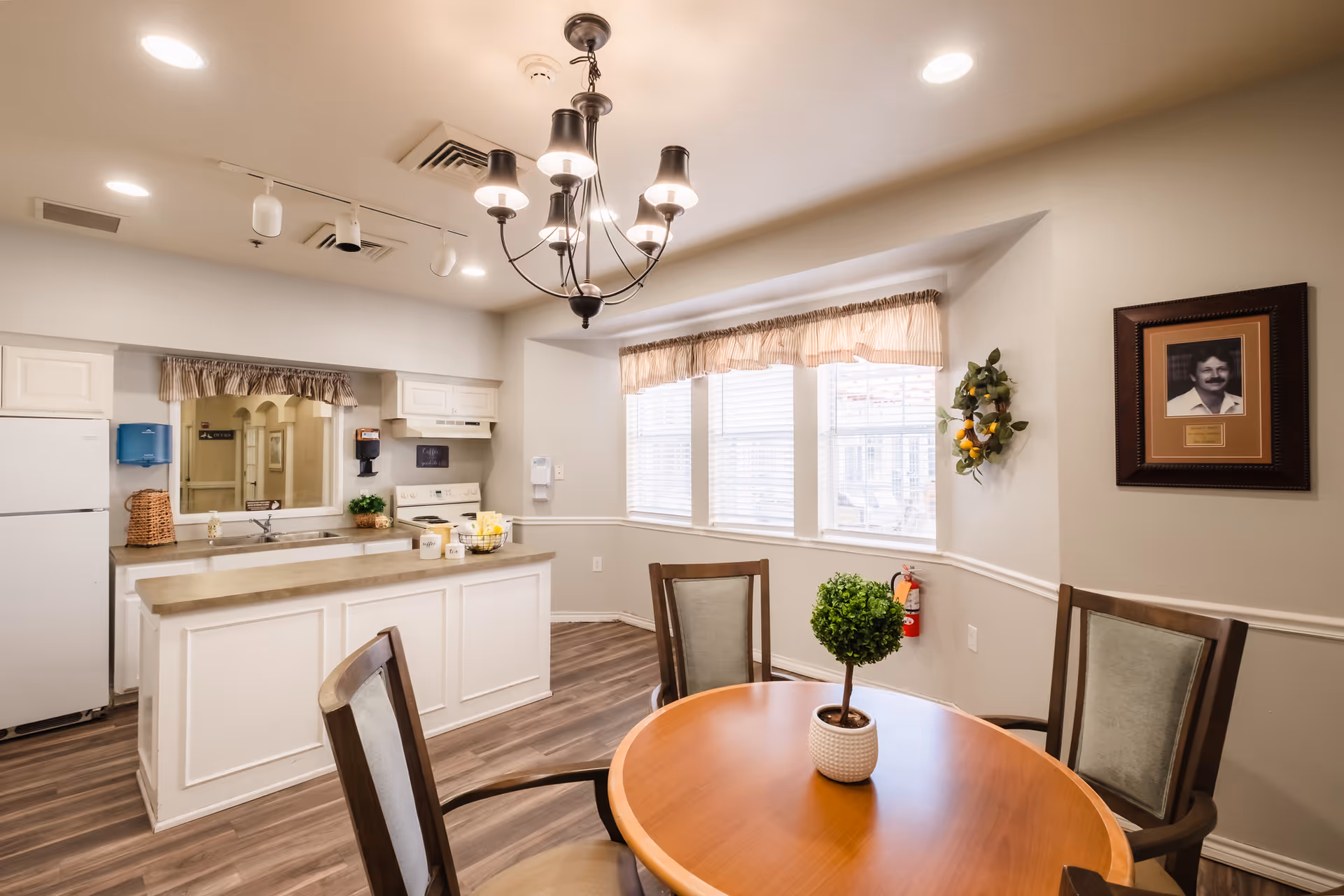Well-lit communal dining area with a round wooden table and chairs, an open kitchen island, chandelier, and large bay window.