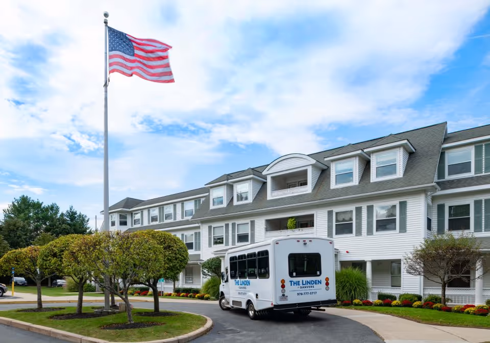Exterior view of a senior living facility with a white multi-story building, neatly trimmed trees, and an American flag on a tall flagpole. A white shuttle bus labeled 'The Linden Danvers' is parked in front of the building.