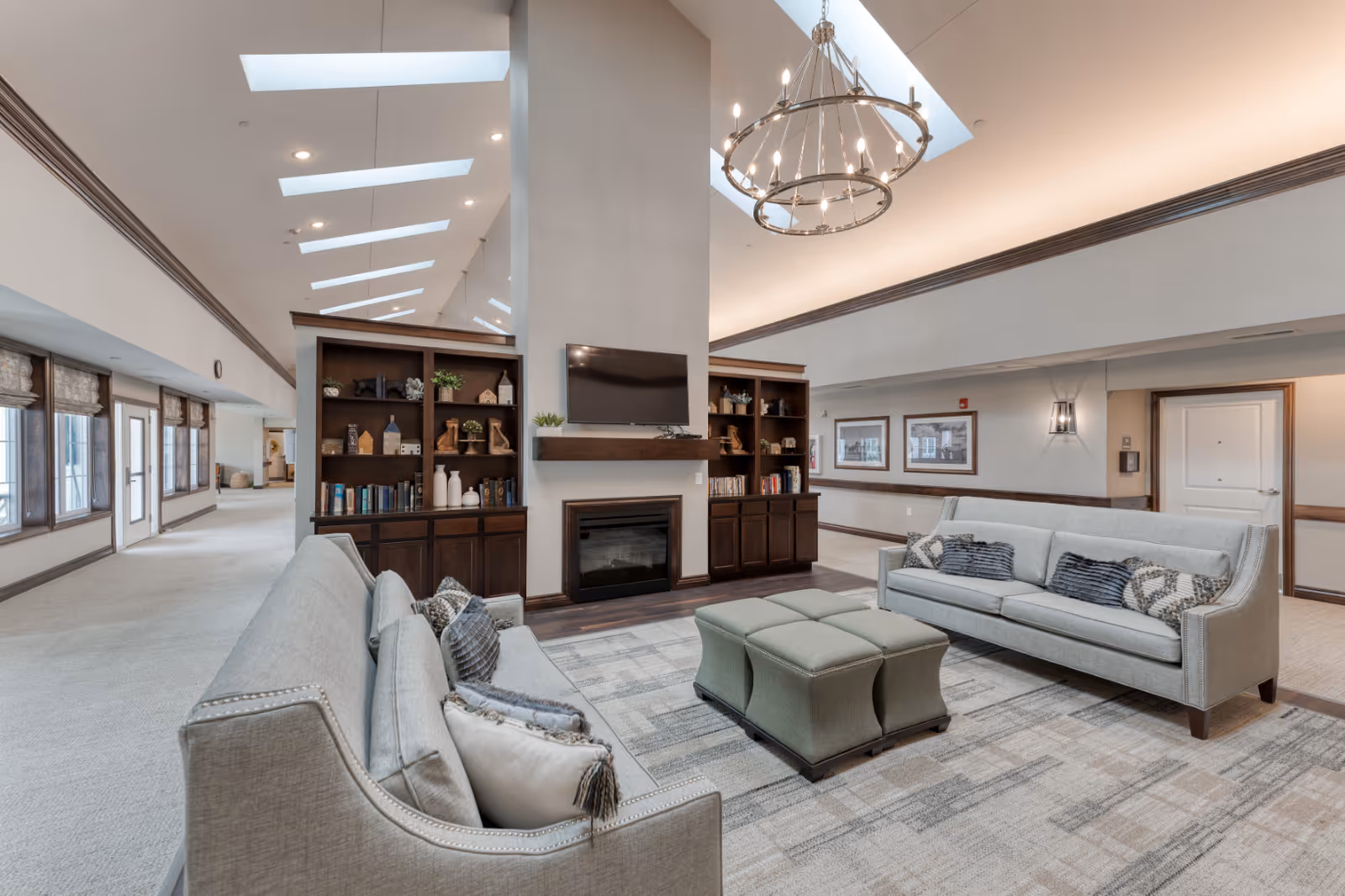 A spacious and well-lit senior living facility common area featuring two light gray sofas with decorative pillows facing each other across a patterned area rug. Between the sofas is a green upholstered ottoman. Behind one sofa is a built-in wooden shelving unit with books, decorative items, and a mounted flat-screen TV above a fireplace. The ceiling is vaulted with skylights and a large chandelier hanging in the center. The walls are painted in neutral tones with framed artwork and wall sconces. Large windows and doors line one side of the room, allowing natural light to enter.