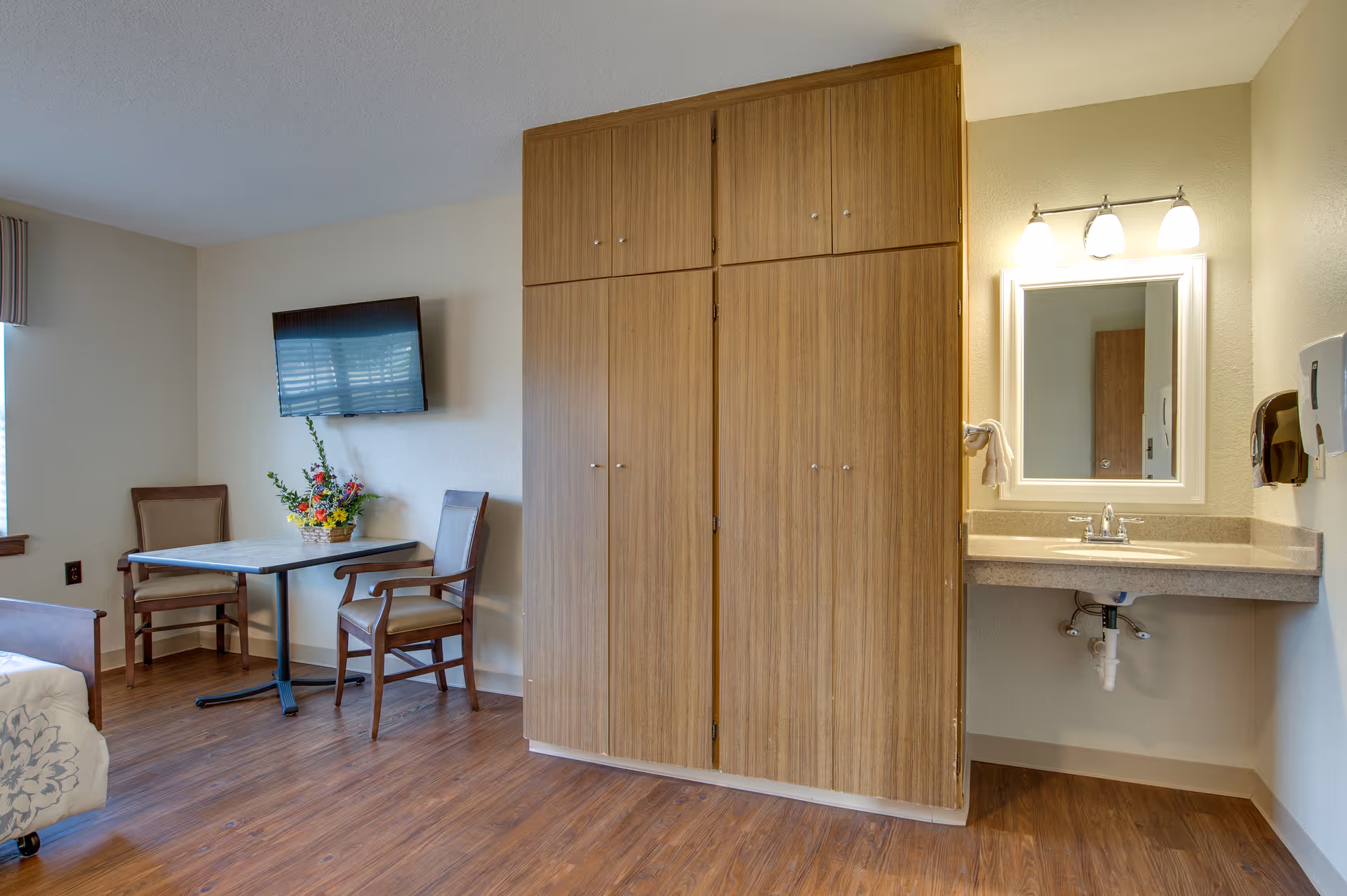 A room with wooden flooring featuring a small table with two chairs, a wall-mounted flat screen TV, a large wooden wardrobe, and a sink with a mirror and three light fixtures above it.