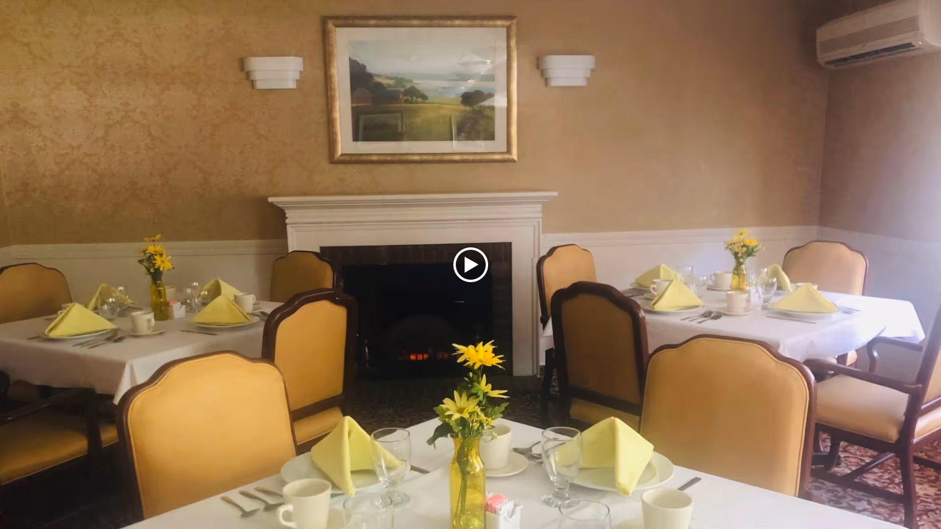 A dining room with three tables set for a meal, each covered with white tablecloths and yellow napkins folded on plates. Each table has a small vase with yellow flowers. The room has beige walls, a fireplace with a framed landscape painting above it, and several upholstered chairs around the tables.