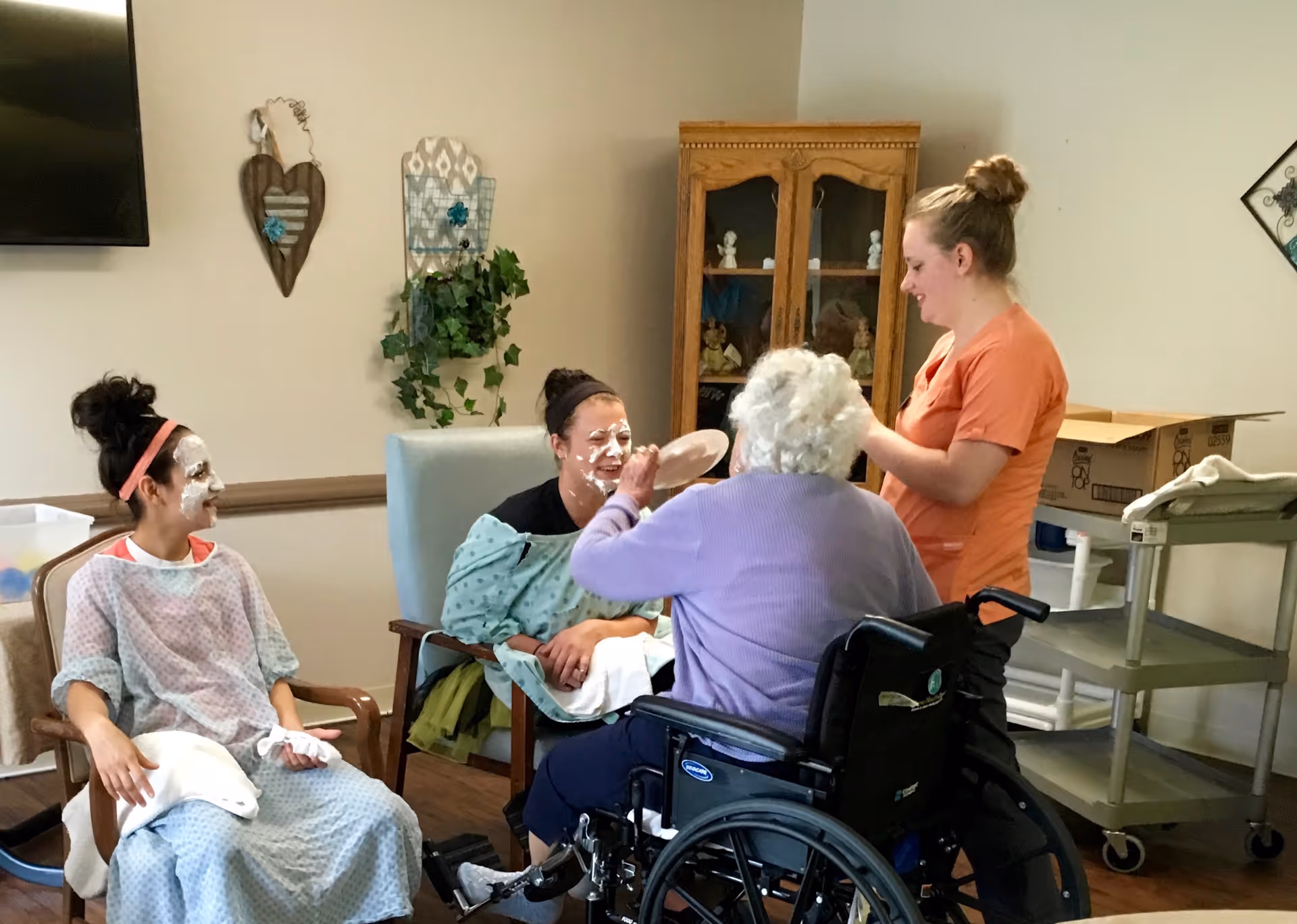 Two young women with face masks sit in chairs while an elderly woman in a wheelchair playfully smears a face mask on one of the young women's faces. A caregiver in orange scrubs stands nearby smiling. The room has light-colored walls, a wooden cabinet with decorative items, and some wall decorations including a heart-shaped piece and a plant.