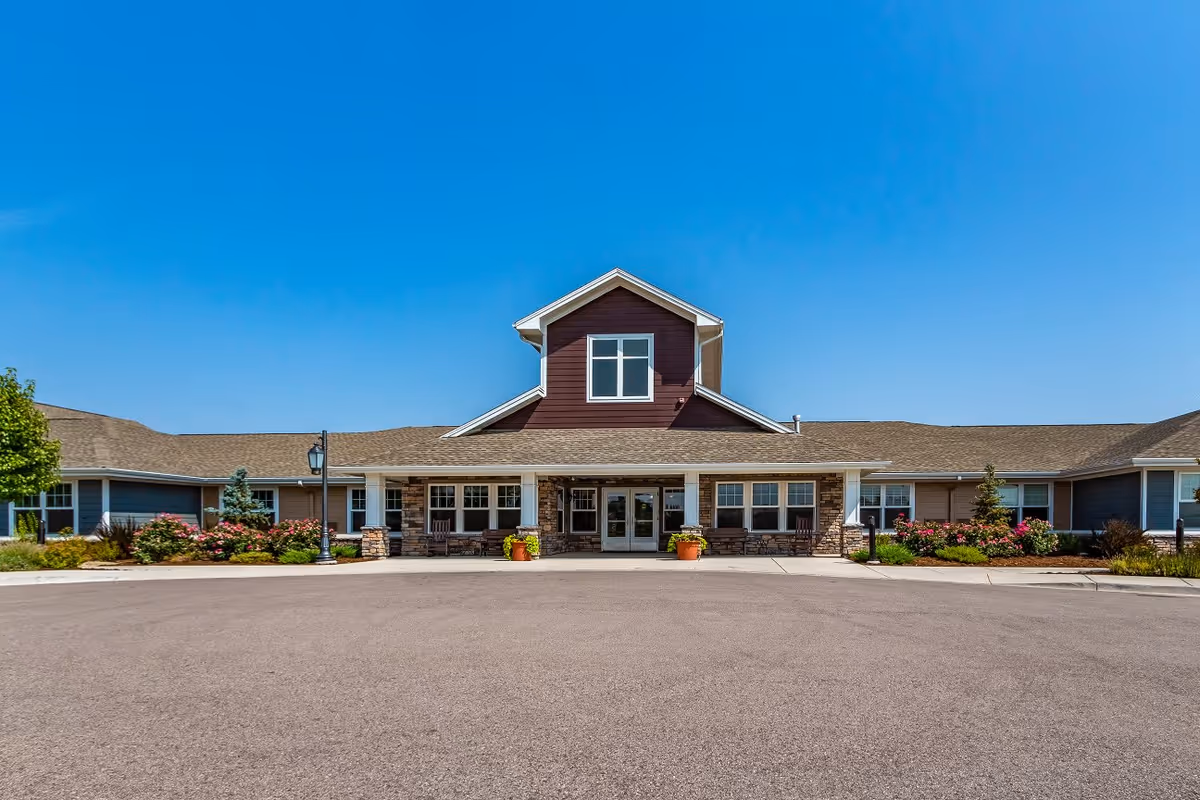 Front exterior view of a single-story senior living facility building with a central entrance, large windows, stone accents, and a peaked roof under a clear blue sky. There are landscaped flower beds and shrubs on either side of the entrance.