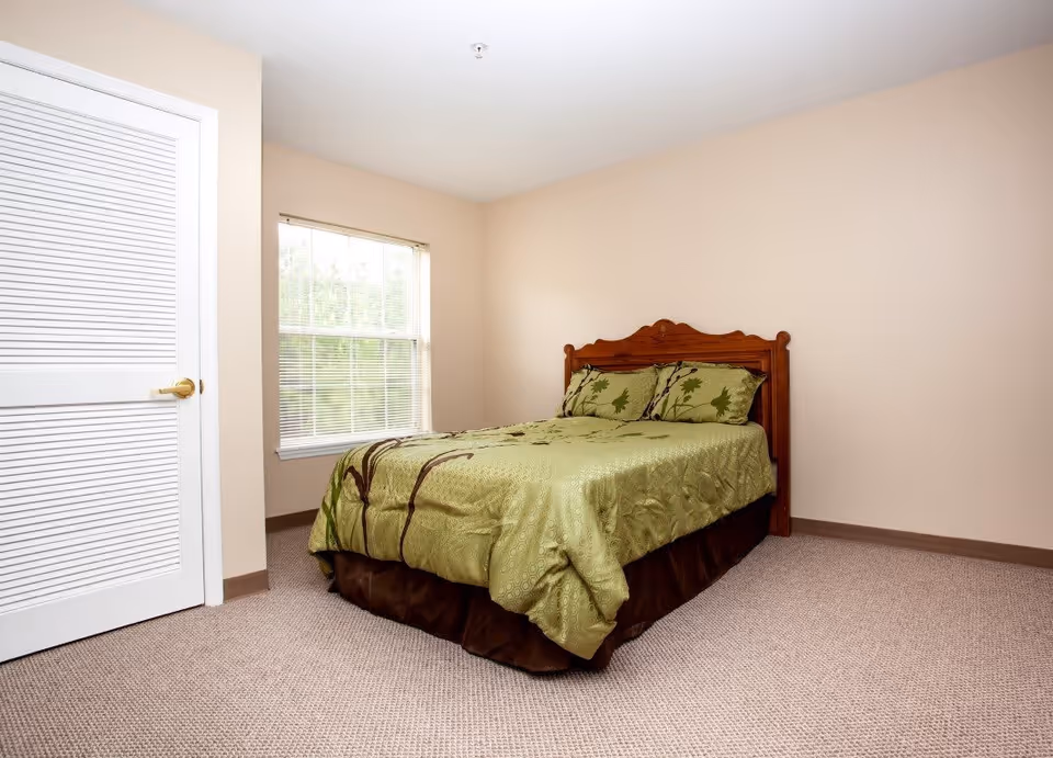 A simple bedroom with a wooden bed frame and a green bedspread with leaf patterns. There is a window with blinds on the left wall and a closed white door with a gold handle next to it. The walls are painted beige and the floor is carpeted.