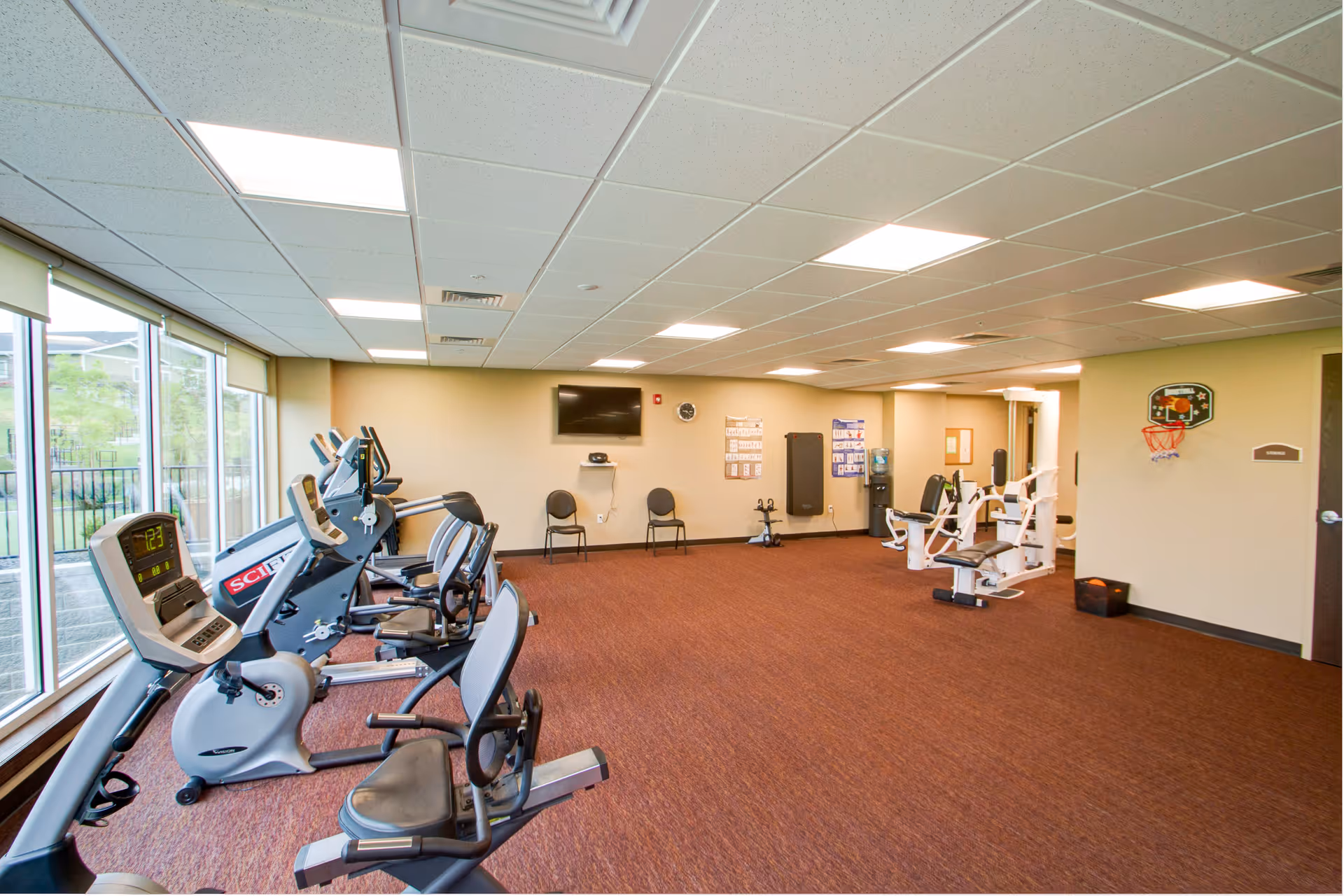 A spacious fitness room with exercise equipment including stationary bikes and weight machines. The room has large windows on one side allowing natural light to enter, a wall-mounted TV, two chairs, and a small basketball hoop on the wall. The floor is carpeted and the ceiling has recessed lighting.