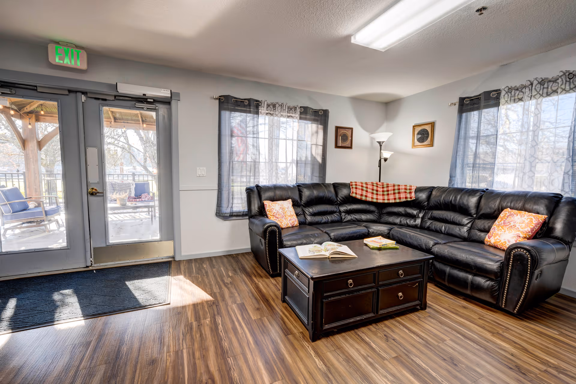 A cozy living room with a large black leather sectional sofa adorned with colorful pillows and a red plaid throw. In front of the sofa is a dark wooden coffee table with books on it. The room has wood flooring, two windows with sheer curtains, a floor lamp, and a glass door leading outside to a patio area with outdoor seating. An illuminated green exit sign is above the door.