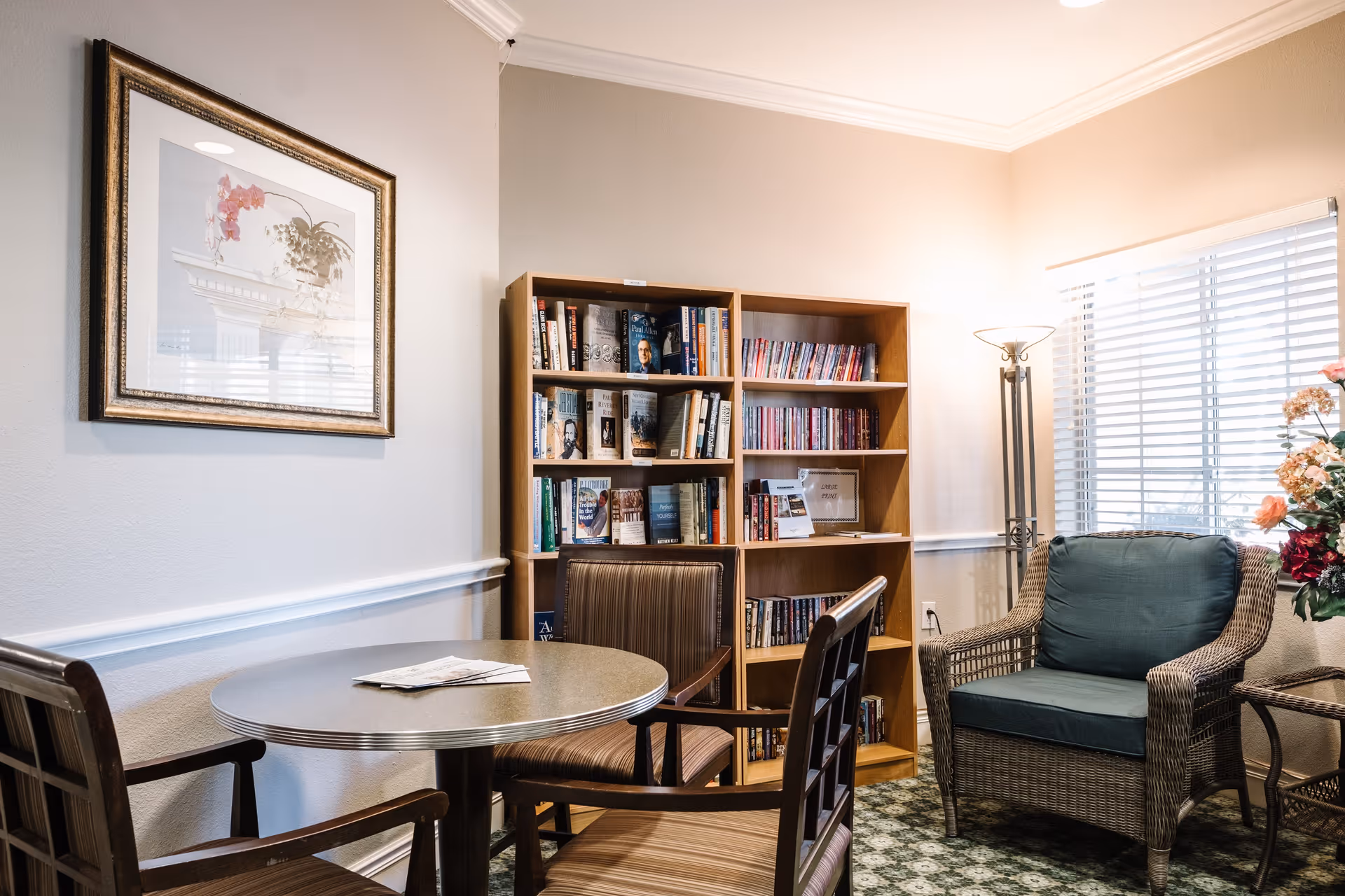 A cozy interior room with a round table surrounded by three wooden chairs, a bookshelf filled with books, a wicker armchair with a green cushion, a floor lamp, a window with blinds, and a framed floral picture on the wall.