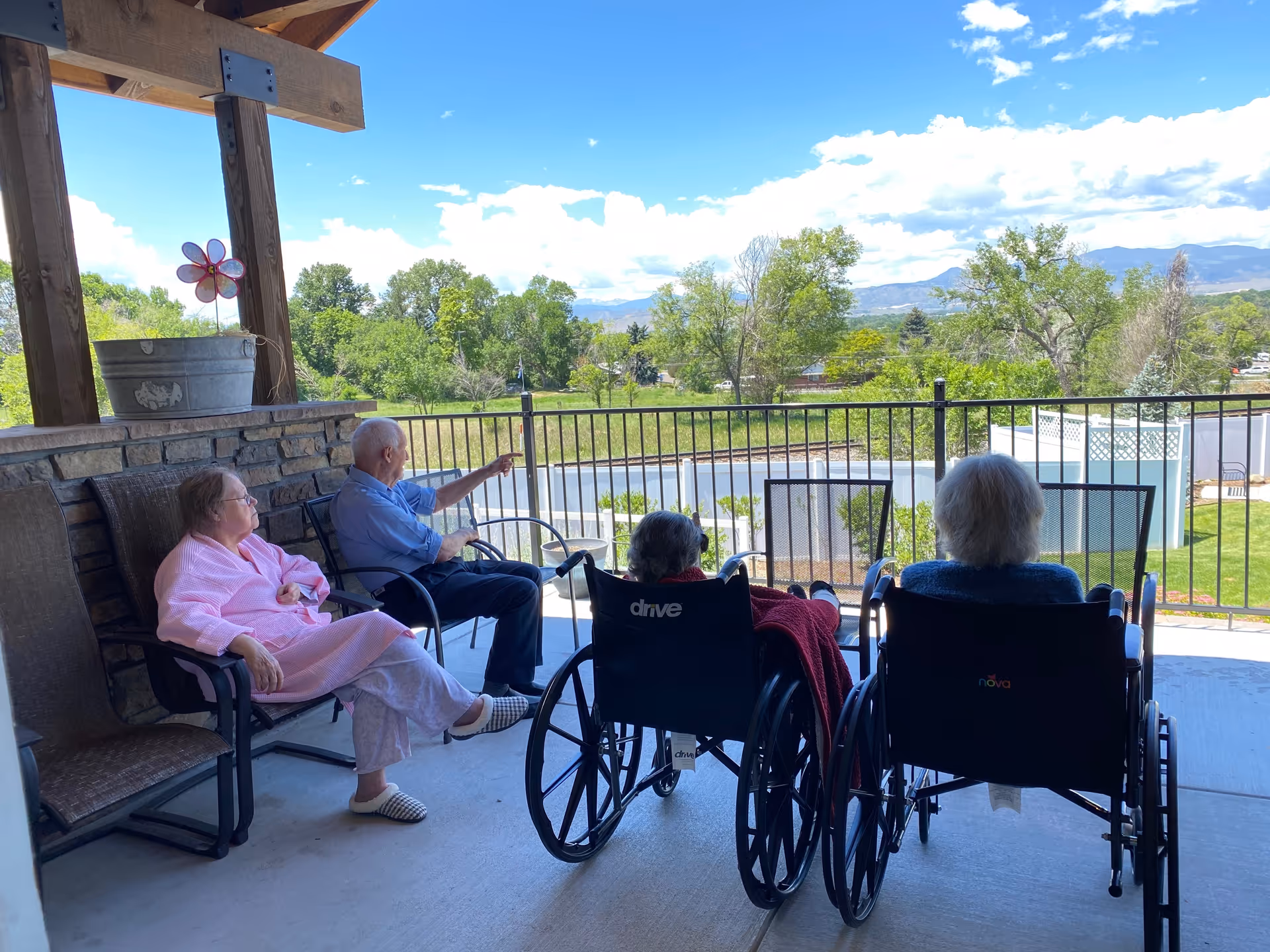 Four elderly individuals sitting on a covered patio overlooking a green outdoor area with trees and mountains in the distance. Two people are seated in wheelchairs facing away from the camera, while the other two sit on chairs. The sky is partly cloudy with blue patches.