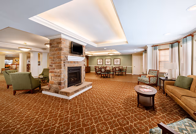 Spacious senior living common area with a central stone fireplace, seating groups, and a dining table in the background.