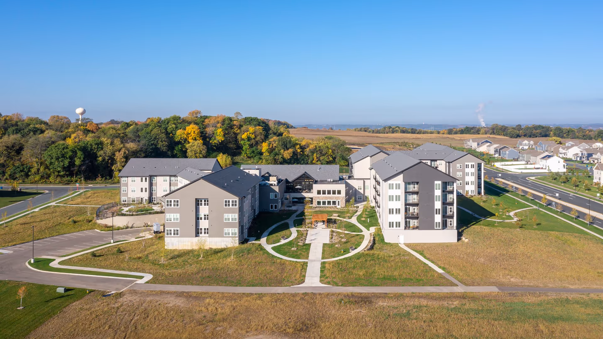 Aerial view of Benedictine Living Community- Shakopee Windermere Way, showing multiple connected residential buildings with gray roofs and beige siding, surrounded by green lawns, paved walkways, and a parking area. Trees with autumn foliage and a clear blue sky are visible in the background.