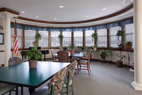 A bright, curved room with large windows covered by blinds and blue valances. Several potted plants are placed on the windowsills and tables. The room contains green tables with floral-patterned chairs and wooden chairs around a central table. An American flag stands in the corner near a piano. The ceiling has recessed lighting and the room features white columns and beige carpeting.