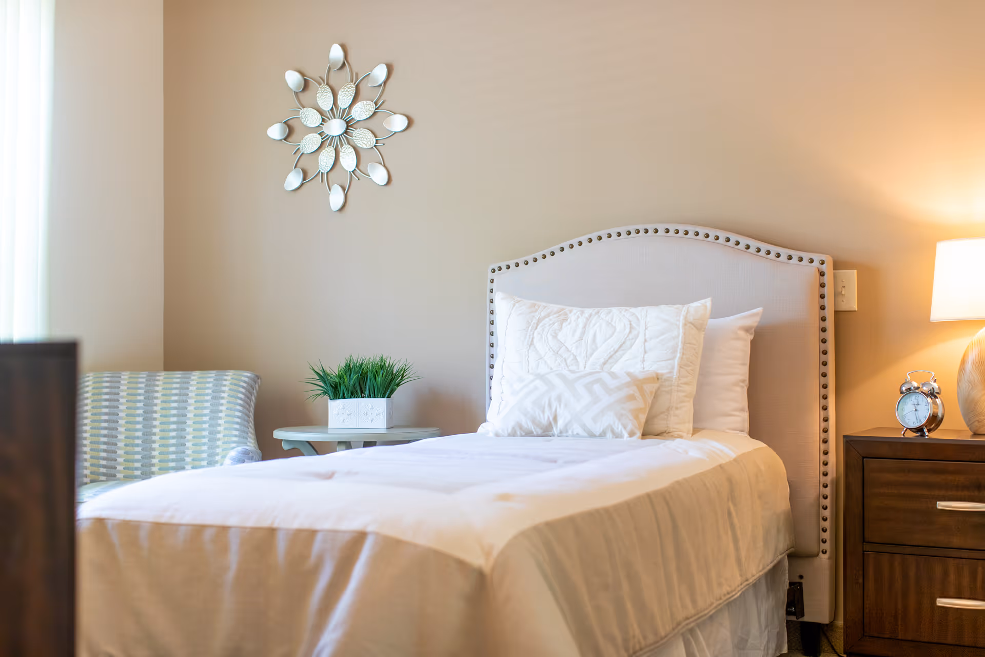 Bright single bed with an upholstered headboard, bedside table with a lamp and alarm clock, a chair, decorative wall mirror and a potted plant.