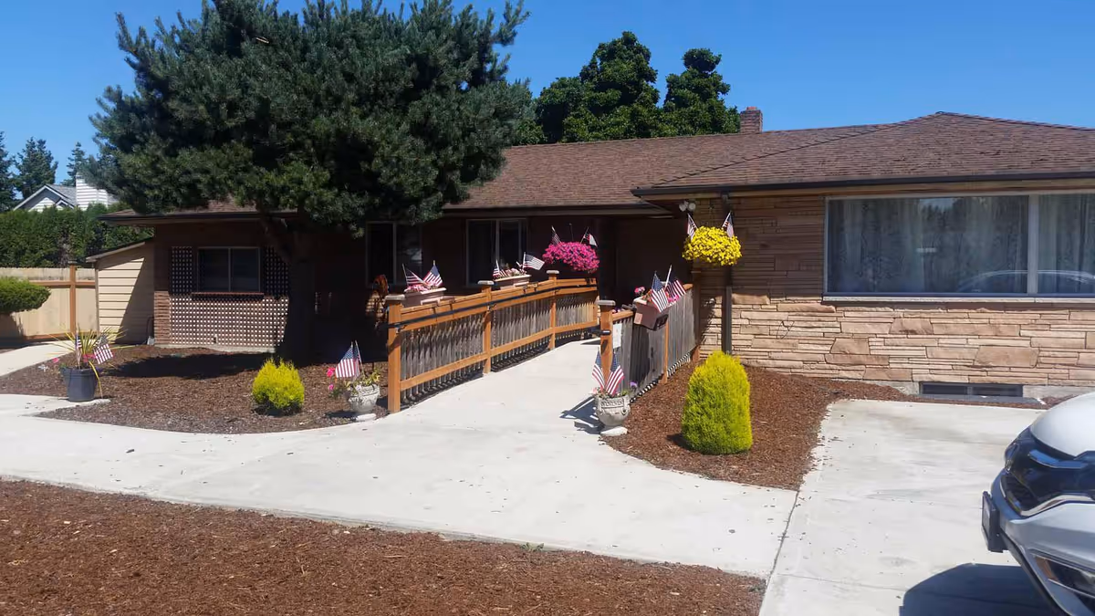 Single-story building front showing an entrance with a wooden accessibility ramp adorned with American flags and hanging flower baskets, plus a driveway.