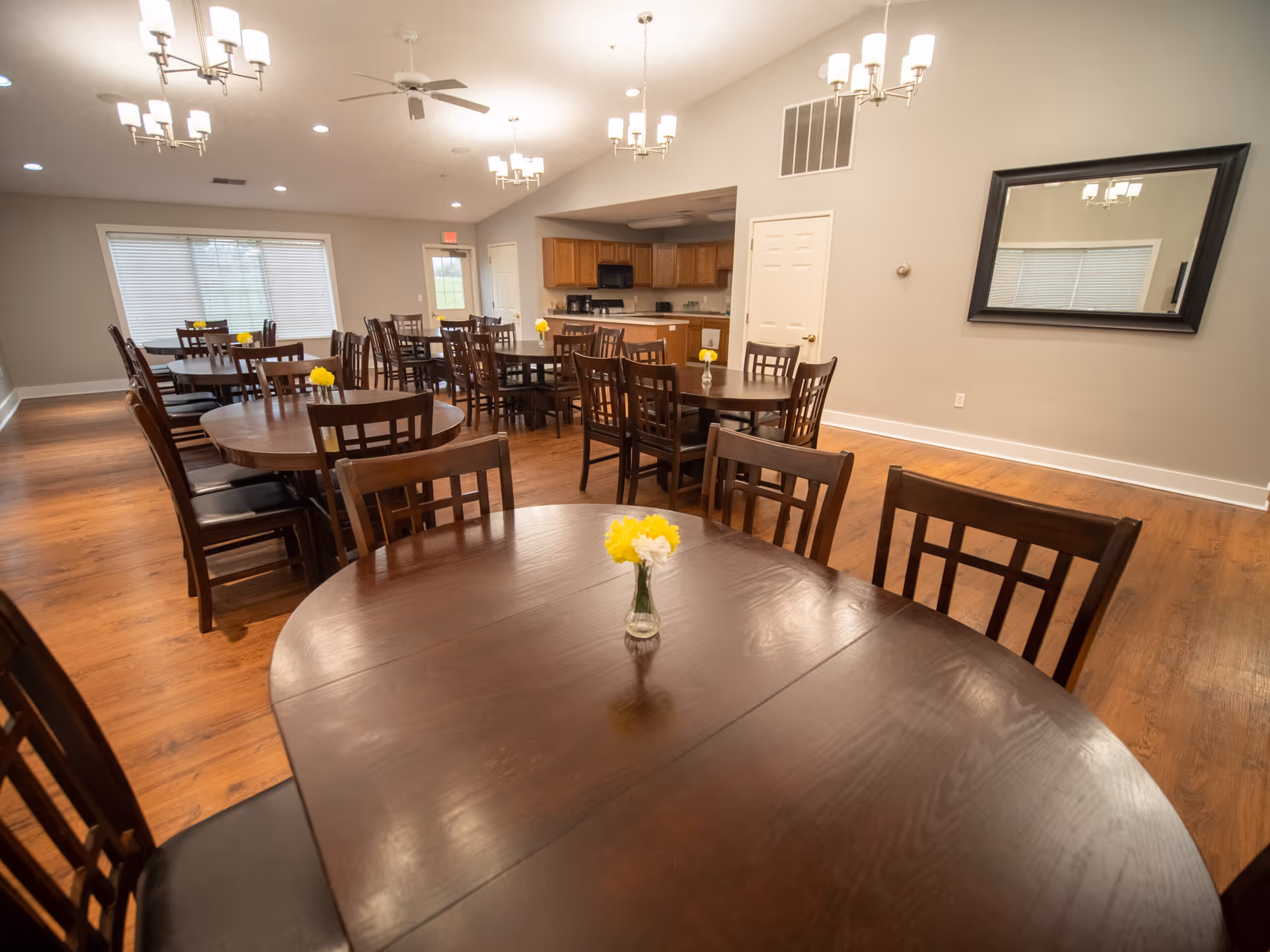 A spacious dining room with multiple round wooden tables and matching chairs. Each table has a small vase with yellow flowers. The room features wooden flooring, beige walls, a large mirror on one wall, and ceiling lights with fans. In the background, there is a kitchen area with wooden cabinets and appliances.