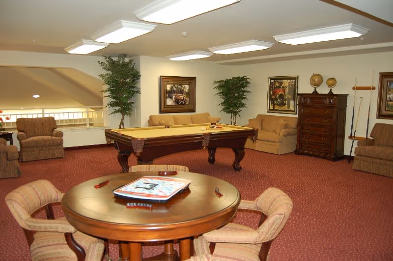 A spacious common area with a pool table in the center, surrounded by beige sofas and armchairs. There is a round wooden table with four upholstered chairs in the foreground, with a board game on top. The room has red carpet flooring, framed artwork on the walls, two tall potted plants, and a wooden chest of drawers with decorative globes and pool cues.