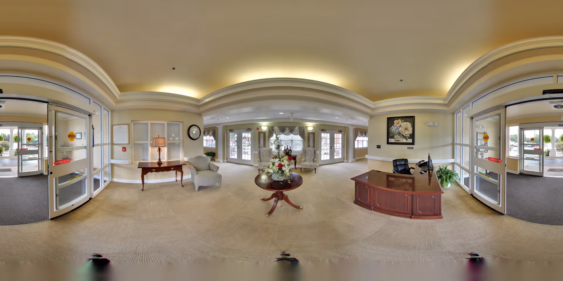A spacious and well-lit senior living facility lobby with beige walls and carpeted floors. The area features a wooden reception desk with a computer and chair, a round table with a floral arrangement in the center, and comfortable armchairs near the windows. There are large glass double doors on both sides of the room allowing natural light to enter. The ceiling has recessed lighting with a soft glow, and decorative elements such as a framed floral painting and a clock are visible on the walls.