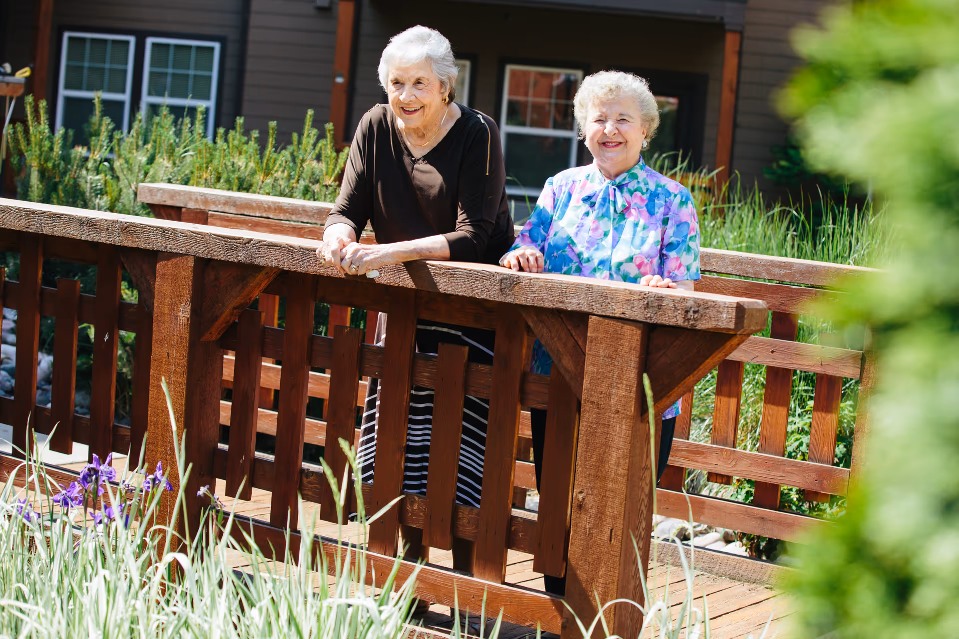 Two elderly women standing and smiling on a wooden bridge surrounded by greenery and flowers, with a building in the background.