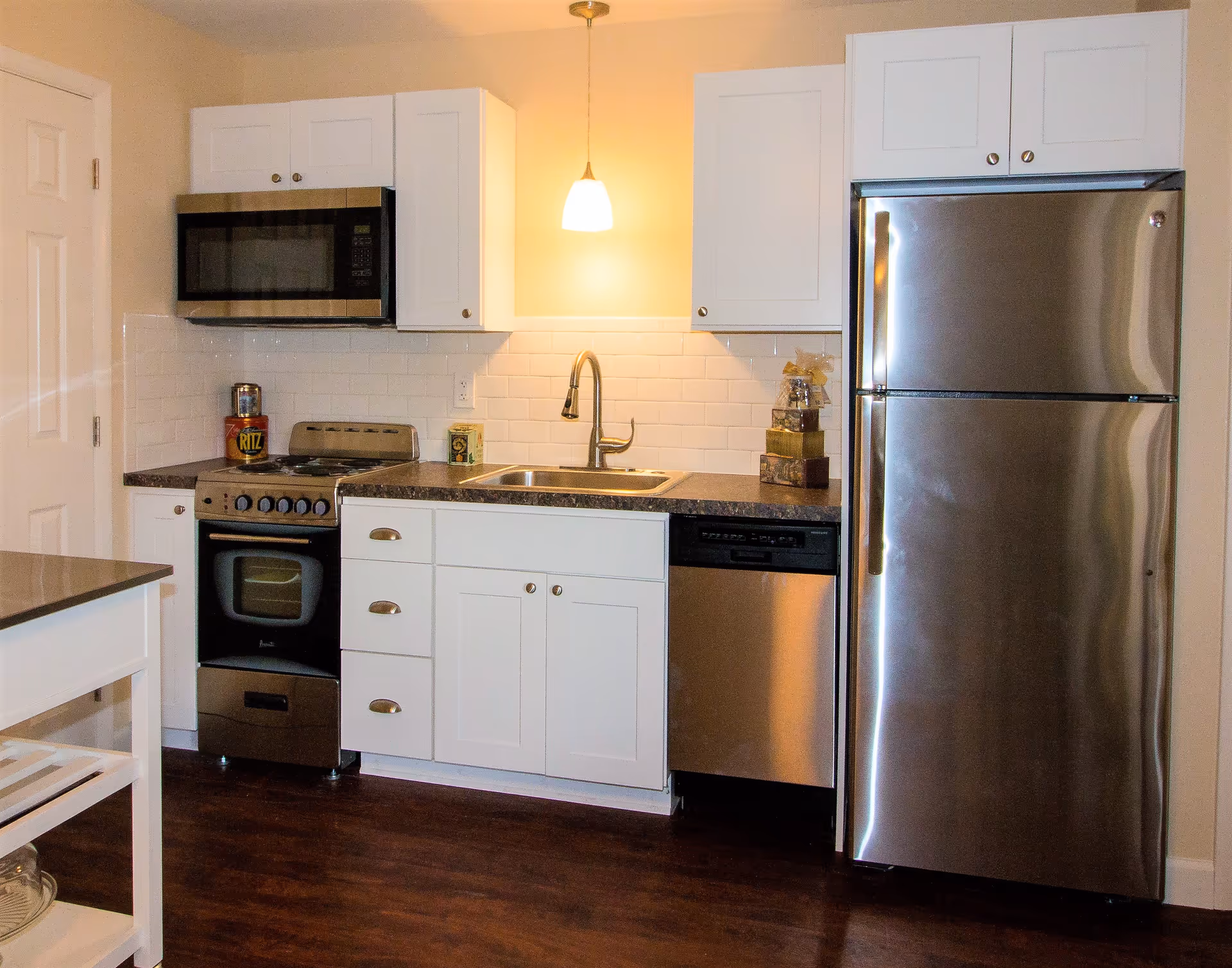 A modern kitchen with white cabinets, a stainless steel refrigerator, dishwasher, oven, and microwave. The kitchen has a dark countertop, a sink with a faucet, and a pendant light hanging above the sink. There are some decorative items on the countertop including a can of Ritz crackers.