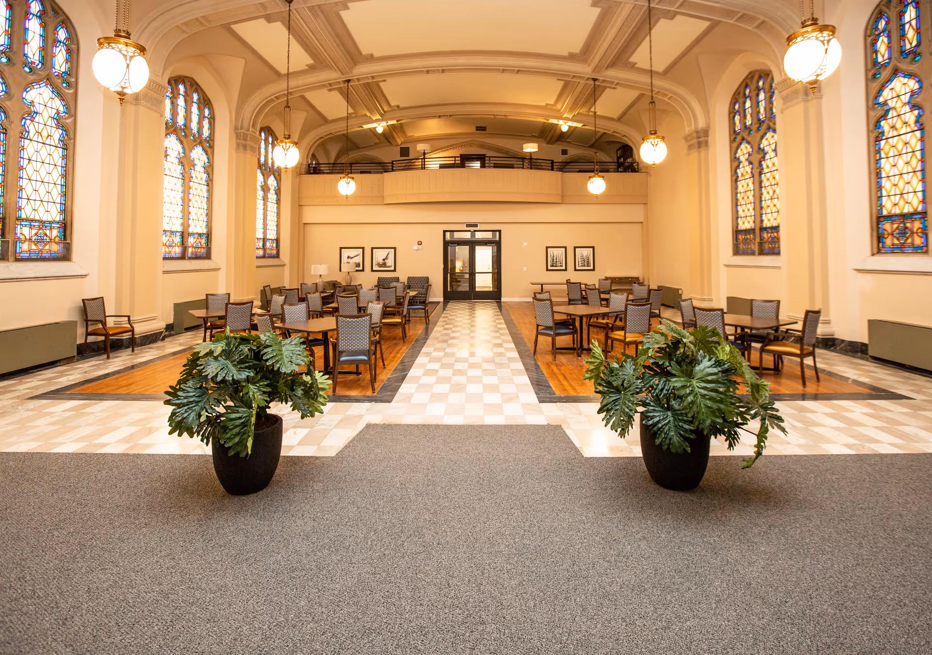 Spacious dining area with multiple tables and chairs arranged neatly on wooden floors, surrounded by tall stained glass windows and hanging pendant lights. Two large potted plants are placed symmetrically in the foreground on a carpeted area, leading to a tiled walkway and double doors at the far end of the room.