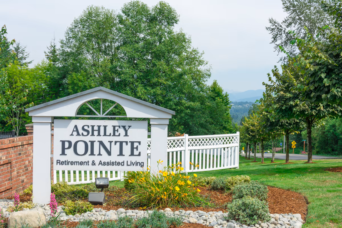 A white sign with black text reading 'Ashley Pointe Retirement & Assisted Living' is displayed outdoors surrounded by landscaping with flowers, bushes, and rocks. Behind the sign is a white fence, trees, and a road leading into a green, wooded area with mountains in the distance under a cloudy sky.