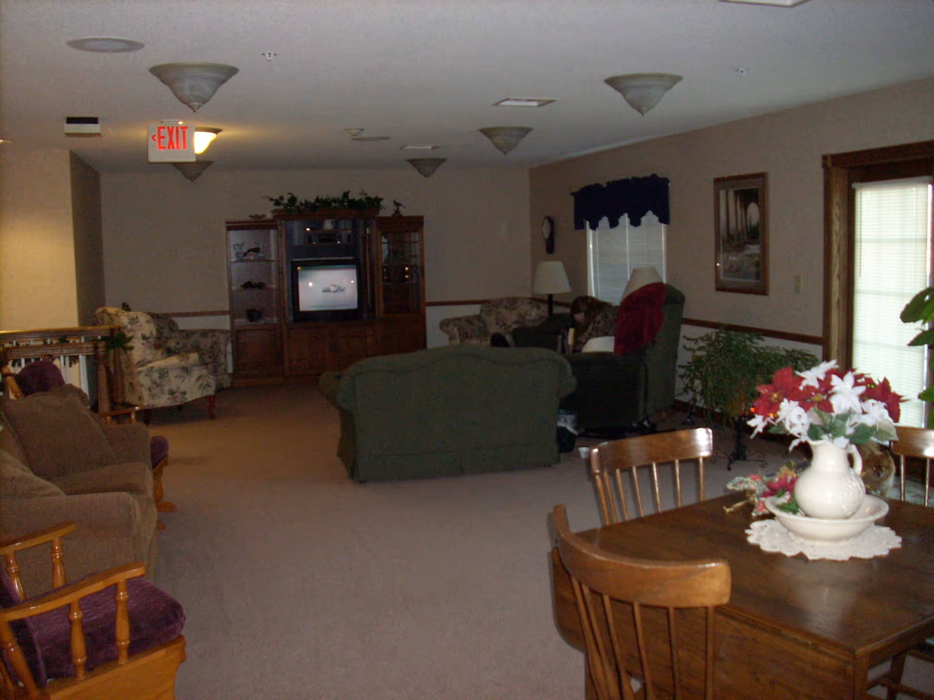 A senior living common room with sofas and armchairs facing a TV cabinet and a dining table with a vase of flowers in the foreground.