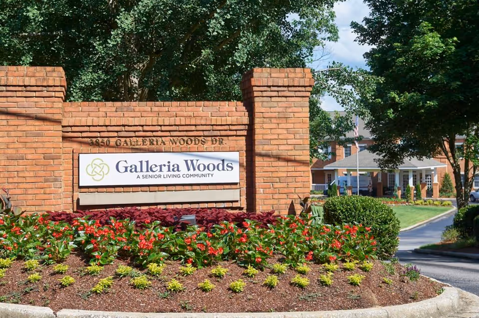 Brick entrance sign for Galleria Woods, a senior living community, surrounded by landscaped flower beds with red and yellow flowers and green bushes, with trees and part of the building visible in the background.