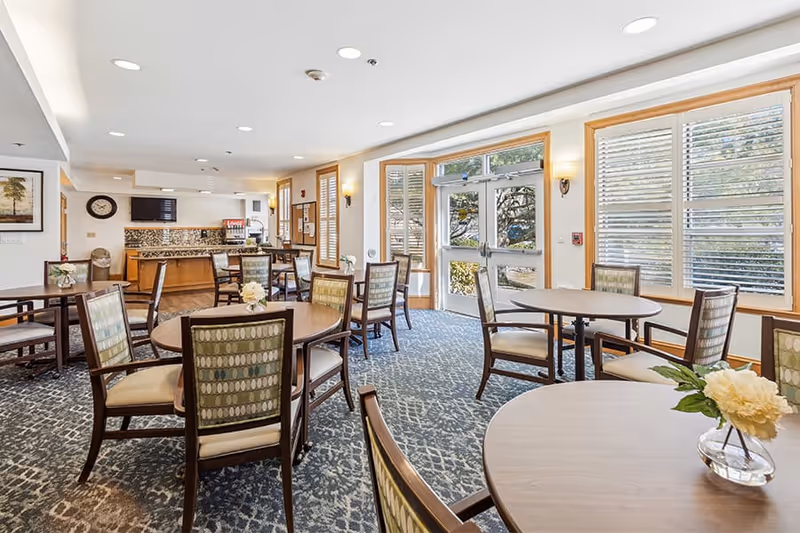 Bright communal dining room with round tables and chairs, patterned carpet, and large windows looking outside.