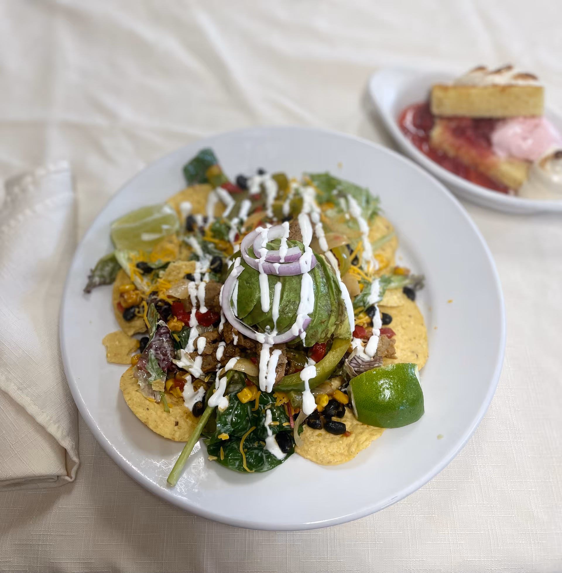 A plate of nachos topped with avocado slices, red onion rings, black beans, corn, lettuce, shredded cheese, and drizzled with white sauce, served with lime wedges on a white tablecloth. In the background, there is a small plate with a slice of cake and a dollop of pink and white cream.