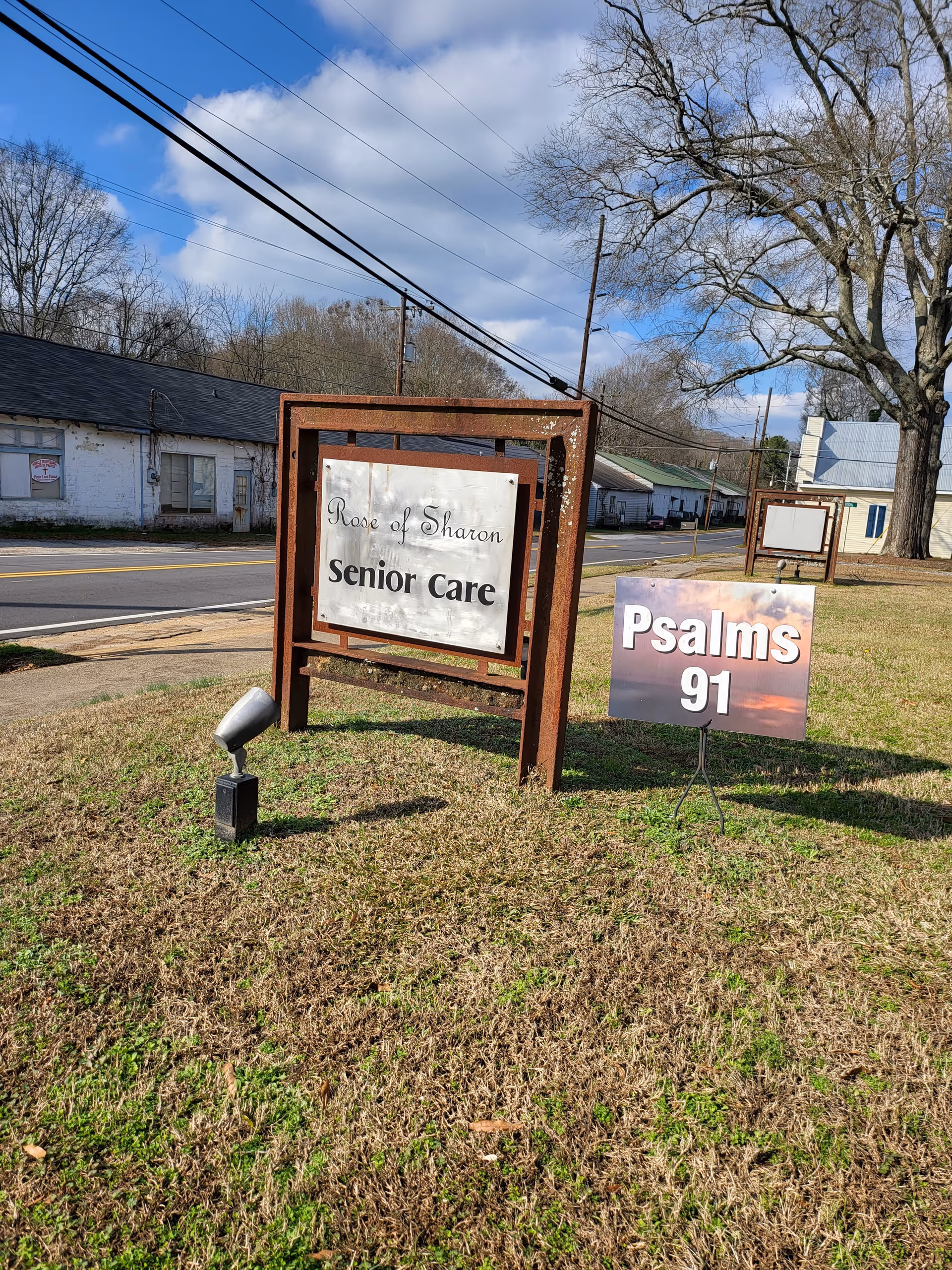 Outdoor view of a roadside sign for Rose of Sharon Senior Care on a grassy area with a smaller sign next to it that reads Psalms 91. There are buildings, a tree, and power lines in the background under a partly cloudy sky.