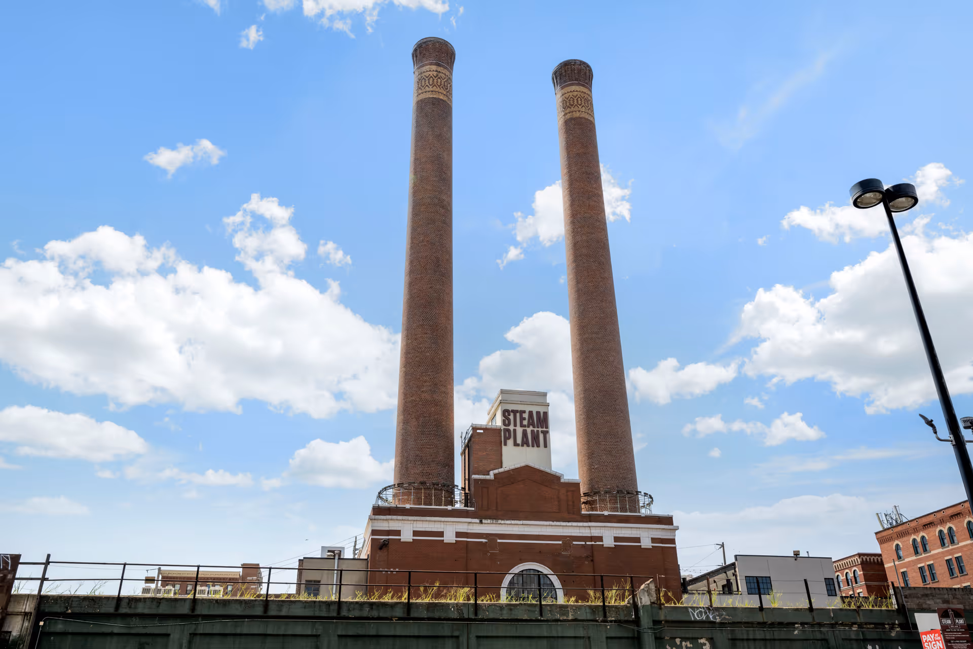 A brick steam plant building with two tall smokestacks under a blue sky with scattered clouds. The building has a sign that reads 'STEAM PLANT'.