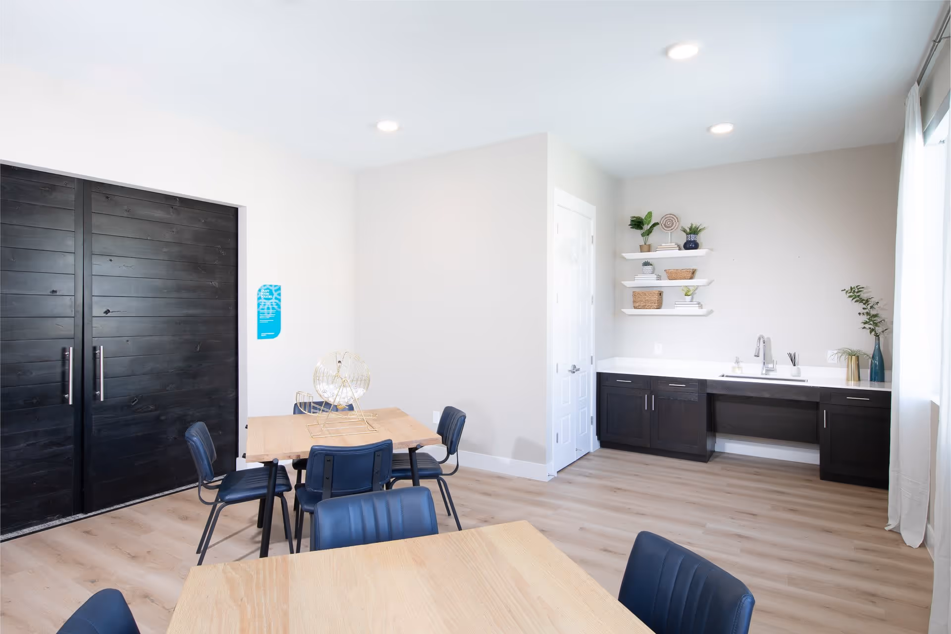 A bright room with light wood flooring and white walls featuring two wooden tables with black chairs around them. One table has a gold bingo cage on it. On the right side, there is a countertop with a sink, dark cabinets underneath, and three white floating shelves with decorative plants and baskets. Large black double doors are on the left side of the room, and a window with white curtains is on the right.