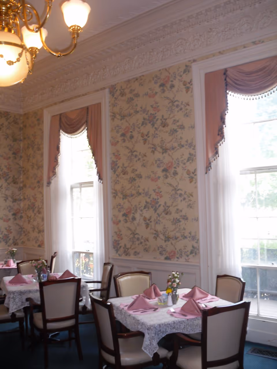 Dining room with small tables set with pink napkins, upholstered chairs, floral wallpaper, tall windows, and a chandelier.
