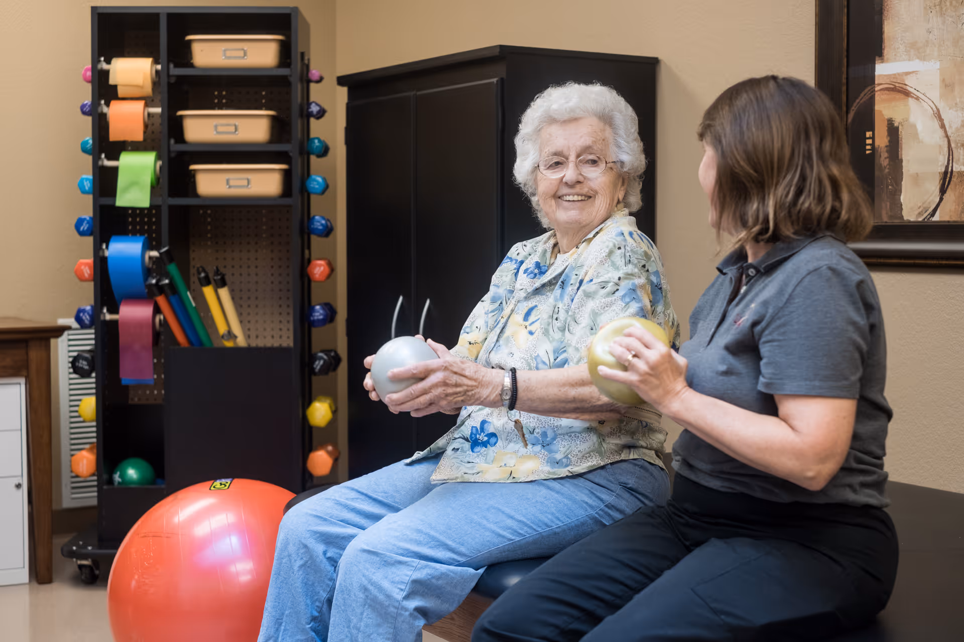 An elderly woman and a younger woman sitting on a bench in a room with exercise equipment. The elderly woman is smiling and holding a silver exercise ball, while the younger woman holds a gold exercise ball. Behind them is a rack with colorful dumbbells and resistance bands.