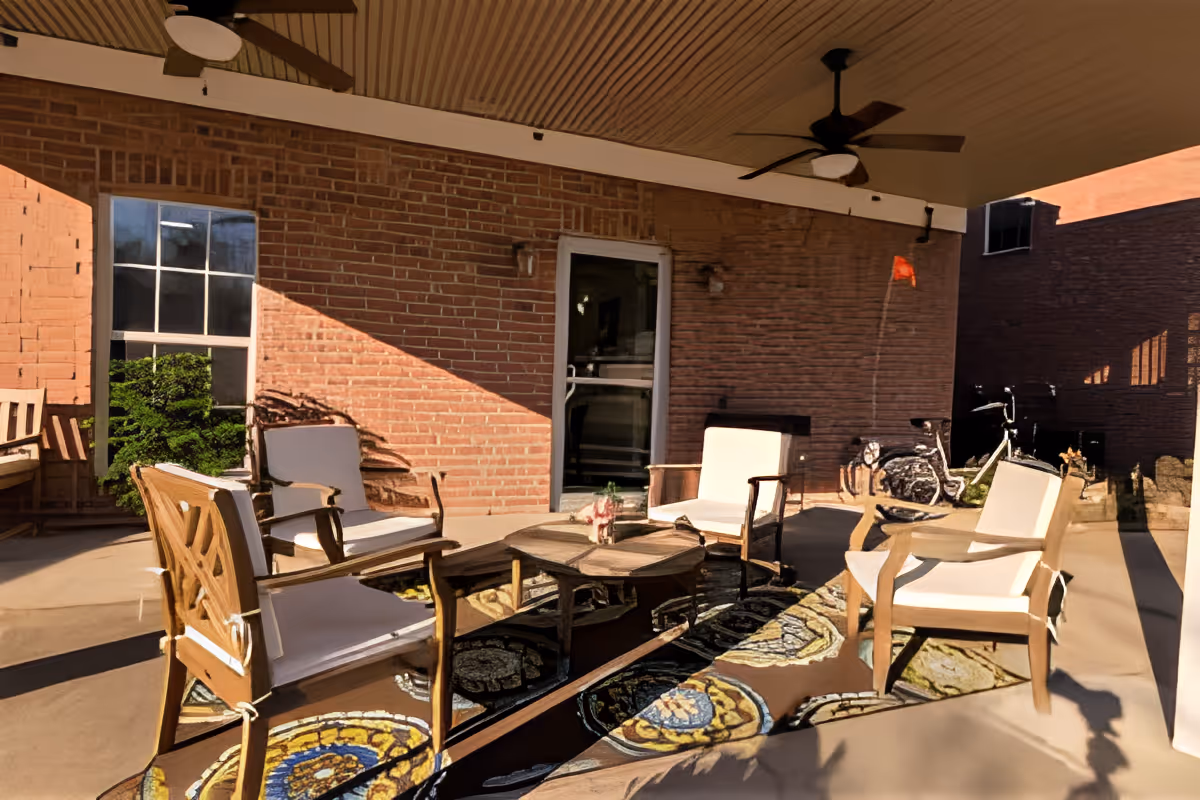 Covered outdoor patio with cushioned chairs and a coffee table arranged under ceiling fans against a brick wall.