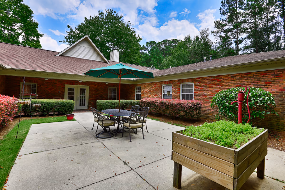 Outdoor patio area at Country Cottages featuring a round metal table with four cushioned chairs and a large green umbrella. The patio is surrounded by a brick building with windows and trimmed bushes. There is a wooden planter box with green plants and a red water pump on the right side. Trees and a partly cloudy sky are visible in the background.