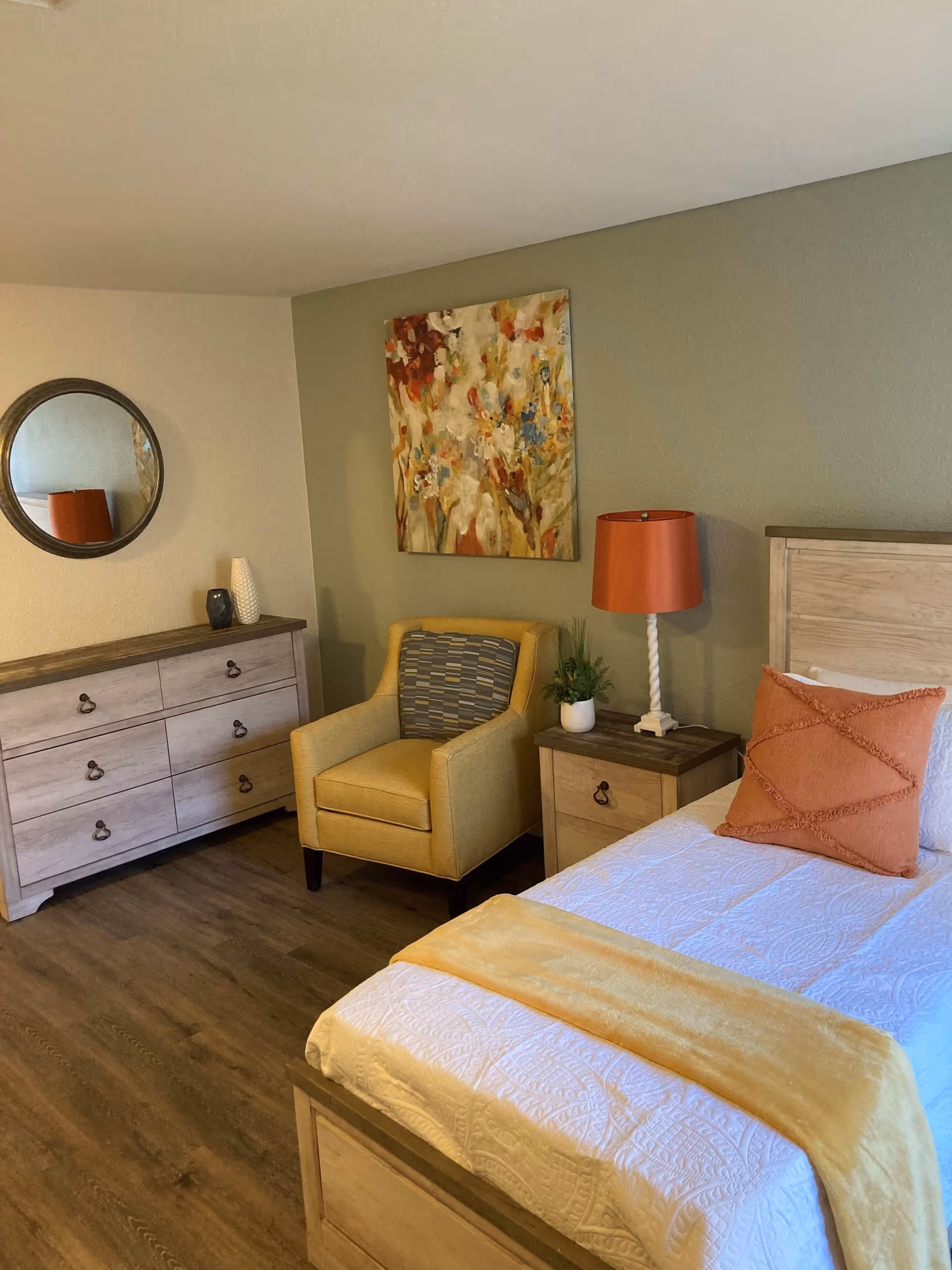 A cozy bedroom featuring a wooden bed with white bedding, an orange decorative pillow, and a yellow throw blanket. Next to the bed is a wooden nightstand with a white lamp with an orange shade and a small potted plant. A yellow armchair with a patterned cushion sits against a green accent wall, above which hangs a colorful abstract painting. On the adjacent wall, there is a wooden dresser with six drawers and a round mirror above it. The room has wood flooring.